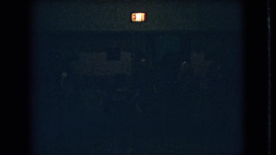 Underexposed 8mm frame from the 1966 Ohio clip with a glowing exit sign above faint human figures in a dark room.
