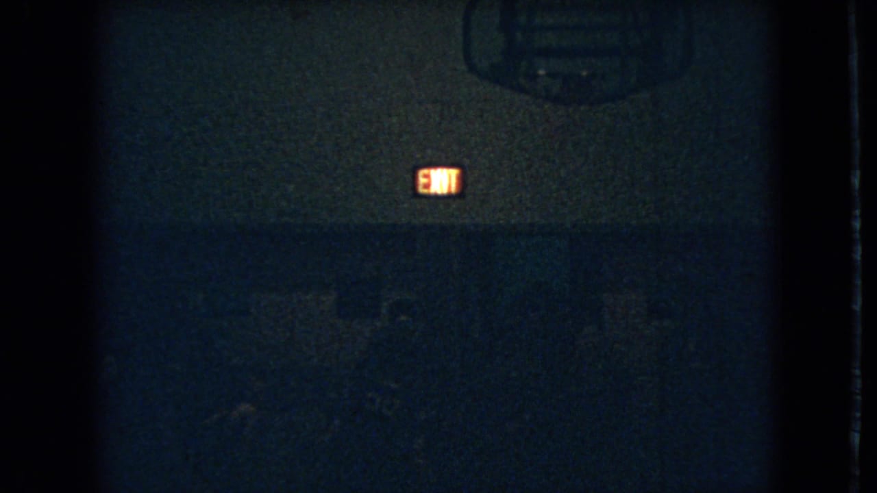 1966 Ohio 8mm frame showing a dim gymnasium interior with a glowing exit sign and barely visible figures in shadow.