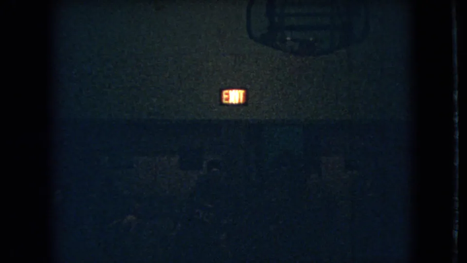 1966 Ohio 8mm frame showing a dim gymnasium interior with a glowing exit sign and barely visible figures in shadow.
