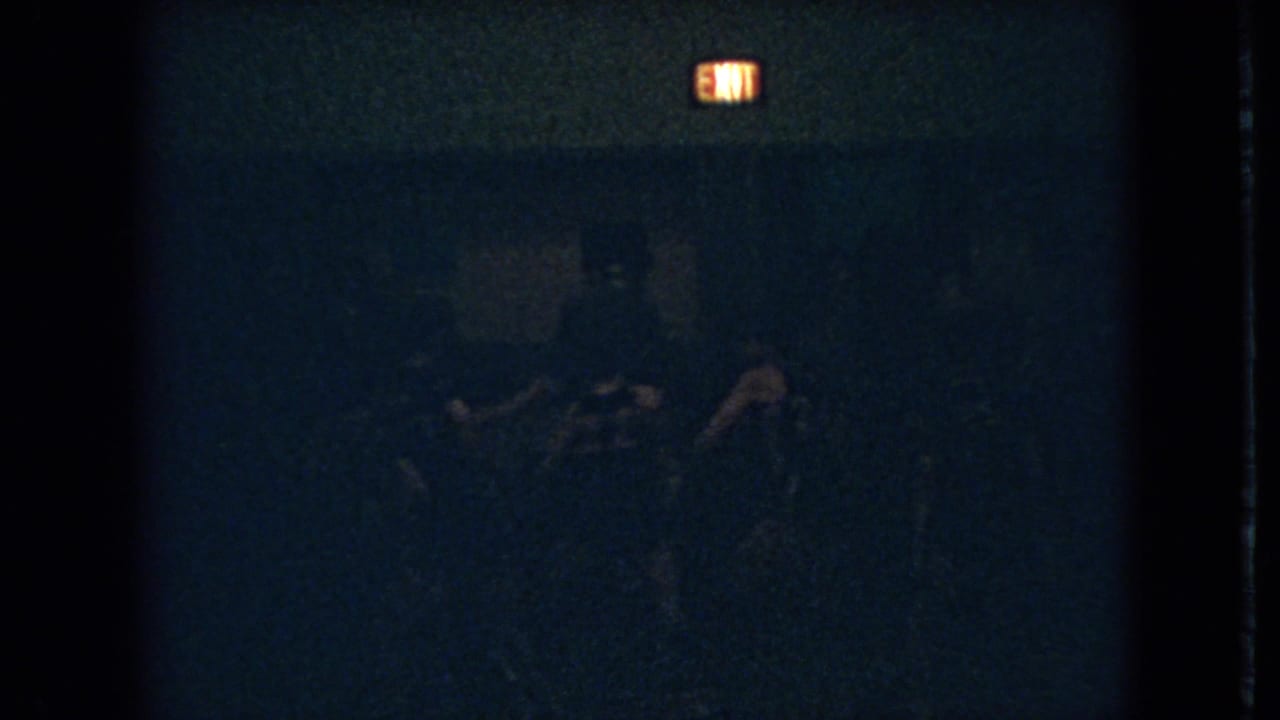 1966 Ohio 8mm frame with a glowing exit sign over faint silhouettes that appear to be players in a dark gymnasium.