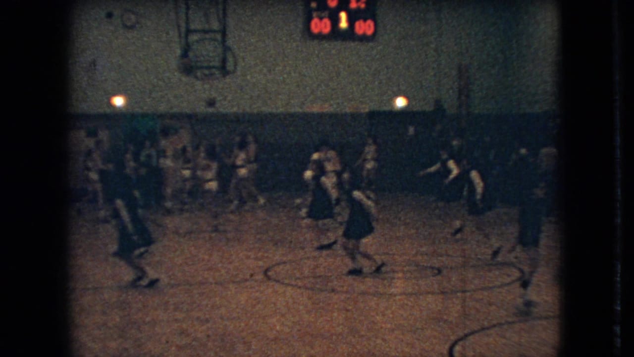 1966 Ohio 8mm home-movie frame showing a distant basketball game in a dim school gym, with players on the court, spectators along the wall, and a scoreboard overhead.