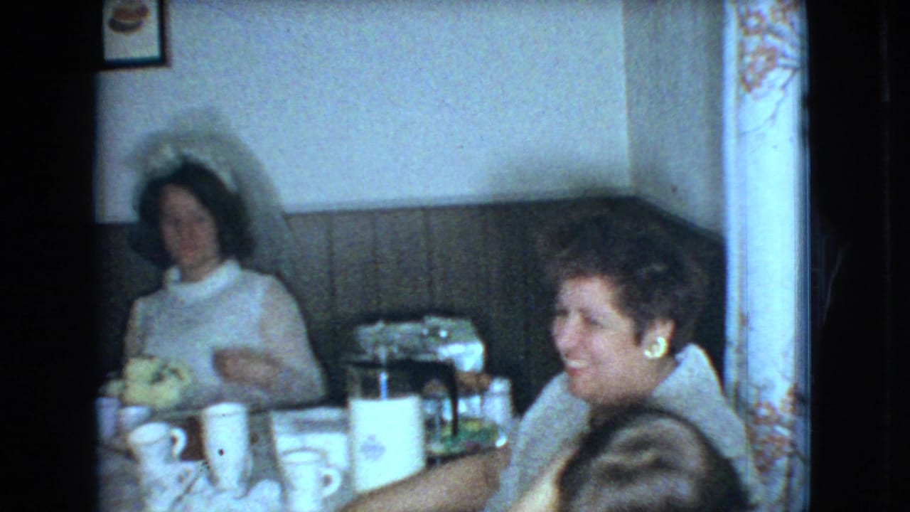 1966 Ohio home-movie frame of a bride in a white dress and veil seated at a reception table with guests, tableware, and a coffee pot in a paneled room.