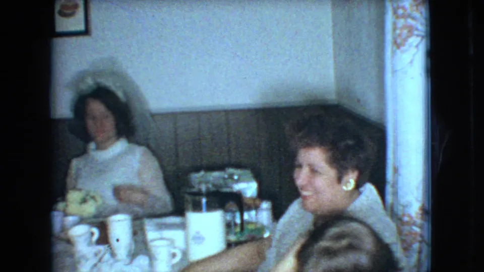 1966 Ohio home-movie frame of a bride in a white dress and veil seated at a reception table with guests, tableware, and a coffee pot in a paneled room.
