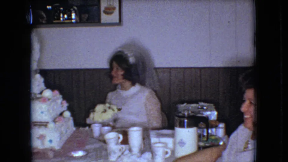 1966 Ohio home-movie frame of the bride seated beside a tiered wedding cake, with cups, a coffee pot, and guests partly visible in a private reception room.