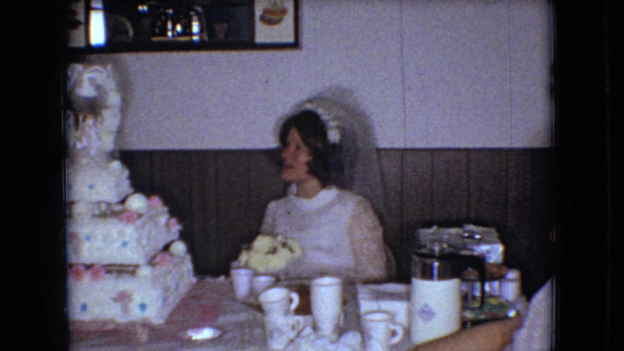 In a 1966 Ohio home-movie frame, the bride sits at a reception table beside a tall decorated wedding cake, with cups, tableware, a thermos, wood paneling, and a wall mirror or framed object behind her.