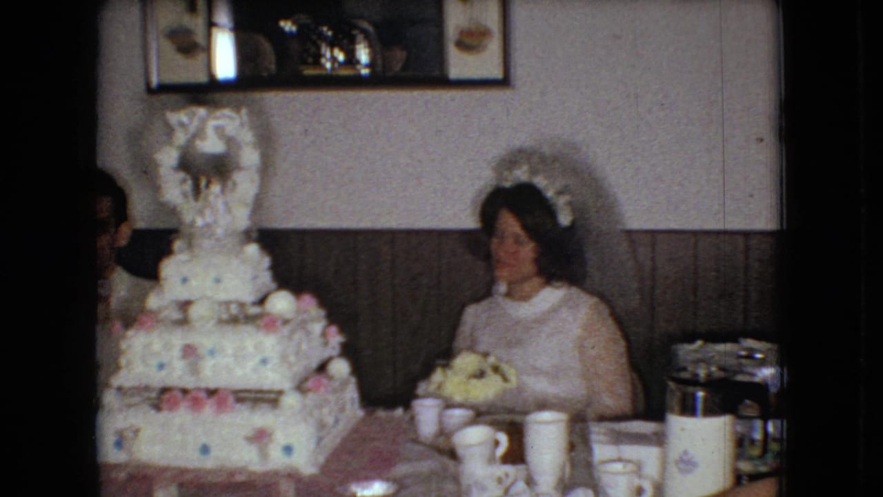 The bride remains seated beside the decorated tiered cake in the 1966 Ohio reception room, with paper cups, a thermos, table settings, wood paneling, and a partial figure near the cake edge.