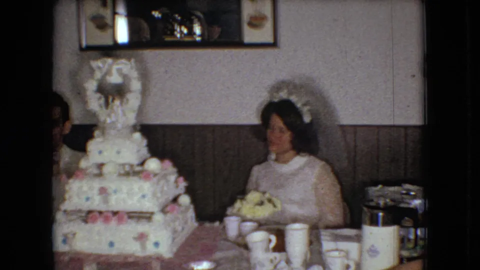 The bride remains seated beside the decorated tiered cake in the 1966 Ohio reception room, with paper cups, a thermos, table settings, wood paneling, and a partial figure near the cake edge.