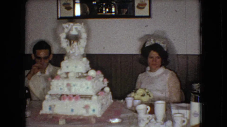 The groom sits at the left and the bride sits at the right behind the decorated wedding cake, with cups, plates, a thermos, and the wood-paneled reception room visible in this 1966 Ohio home-movie frame.