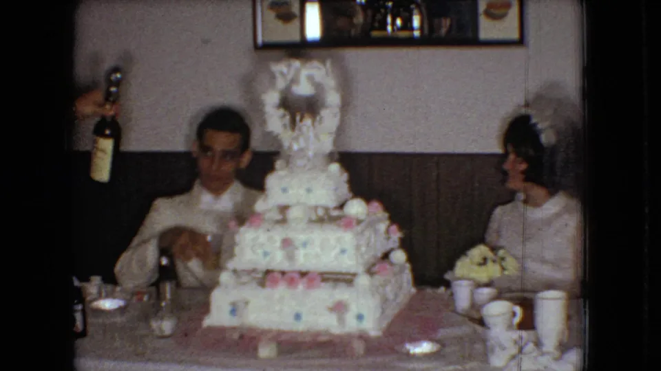 The groom and bride sit at the reception table behind the decorated wedding cake while a hand at the left edge holds a bottle, with cups, tableware, and the wood-paneled interior visible in the 1966 Ohio footage.