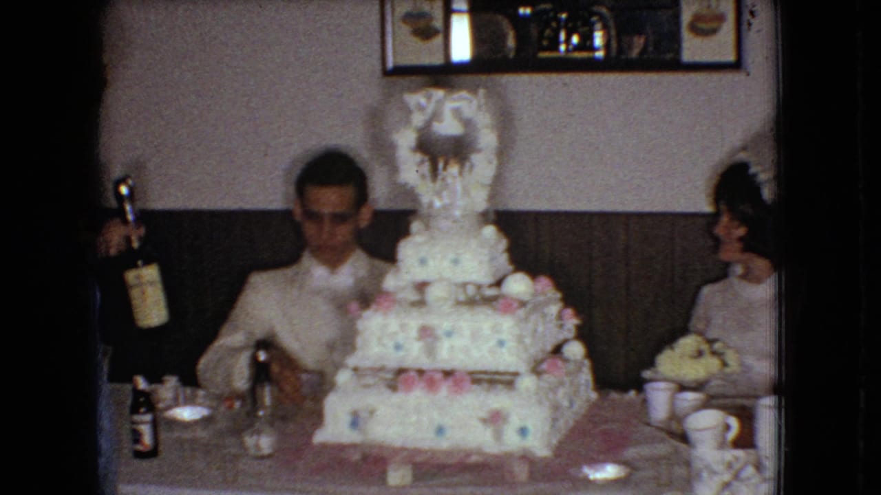 1966 Ohio home-movie frame showing a groom in a white tuxedo jacket seated at a reception table behind a large decorated wedding cake while a nearby guest holds a bottle.