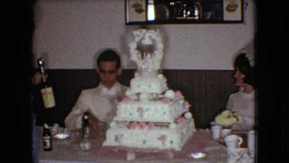 1966 Ohio home-movie frame showing a groom in a white tuxedo jacket seated at a reception table behind a large decorated wedding cake while a nearby guest holds a bottle.