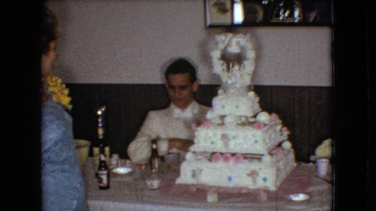 Groom sits at the reception table with the tiered cake in front and a woman partly visible nearby.