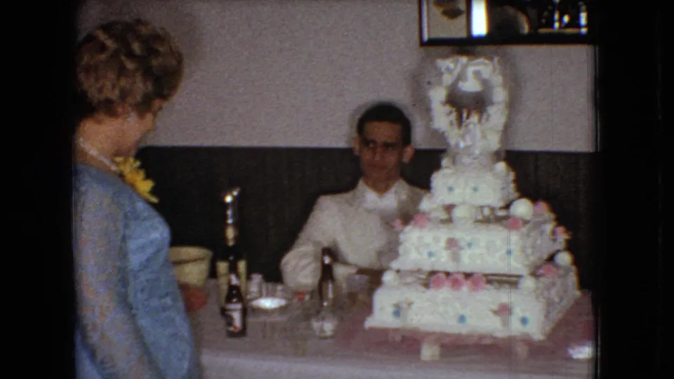 1966 Ohio home-movie frame showing a woman in a light blue dress with a yellow corsage standing beside the reception table as the groom sits behind the tiered wedding cake.