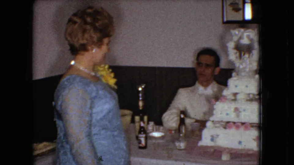 1966 Ohio reception frame with a woman in a light blue dress and corsage standing close to the decorated tiered cake while the groom remains seated at the table.
