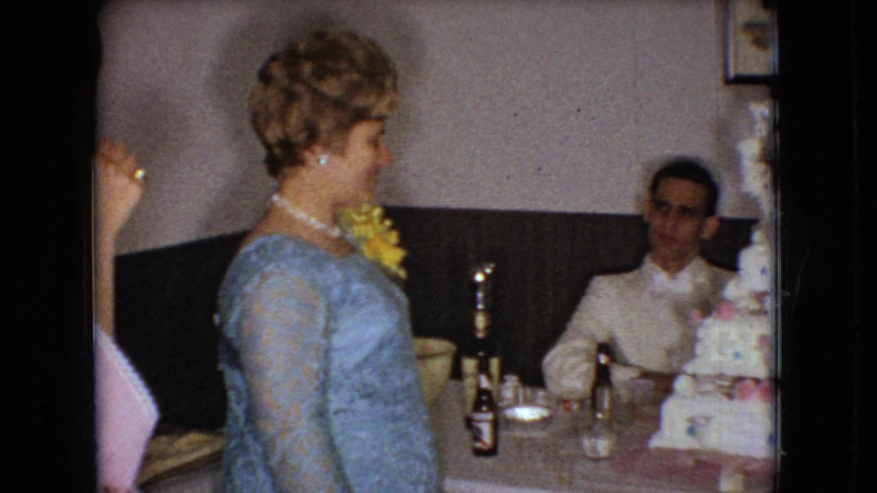 In 1966 Ohio home-movie footage, a woman in a blue lace dress with a yellow corsage and pearl necklace stands near a table with a seated man, bottles, glasses, and part of a tiered cake visible.