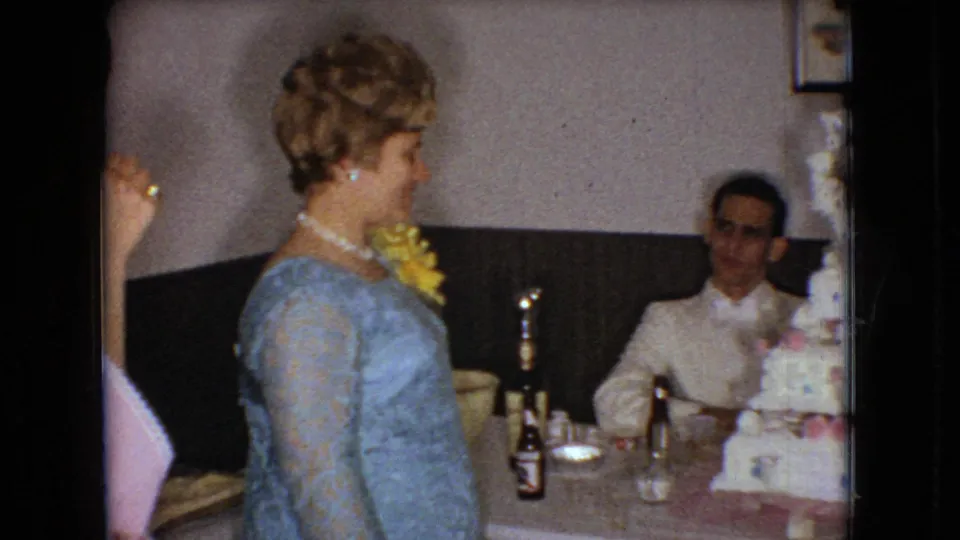 In 1966 Ohio home-movie footage, a woman in a blue lace dress with a yellow corsage and pearl necklace stands near a table with a seated man, bottles, glasses, and part of a tiered cake visible.