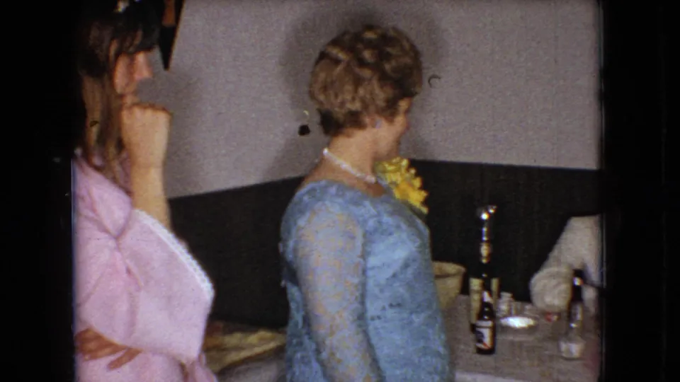 In the indoor 1966 Ohio home-movie frame, a woman in pink appears near the camera while the woman in the blue lace dress remains beside the table with a beer bottle, glasses, and a partially visible seated guest.
