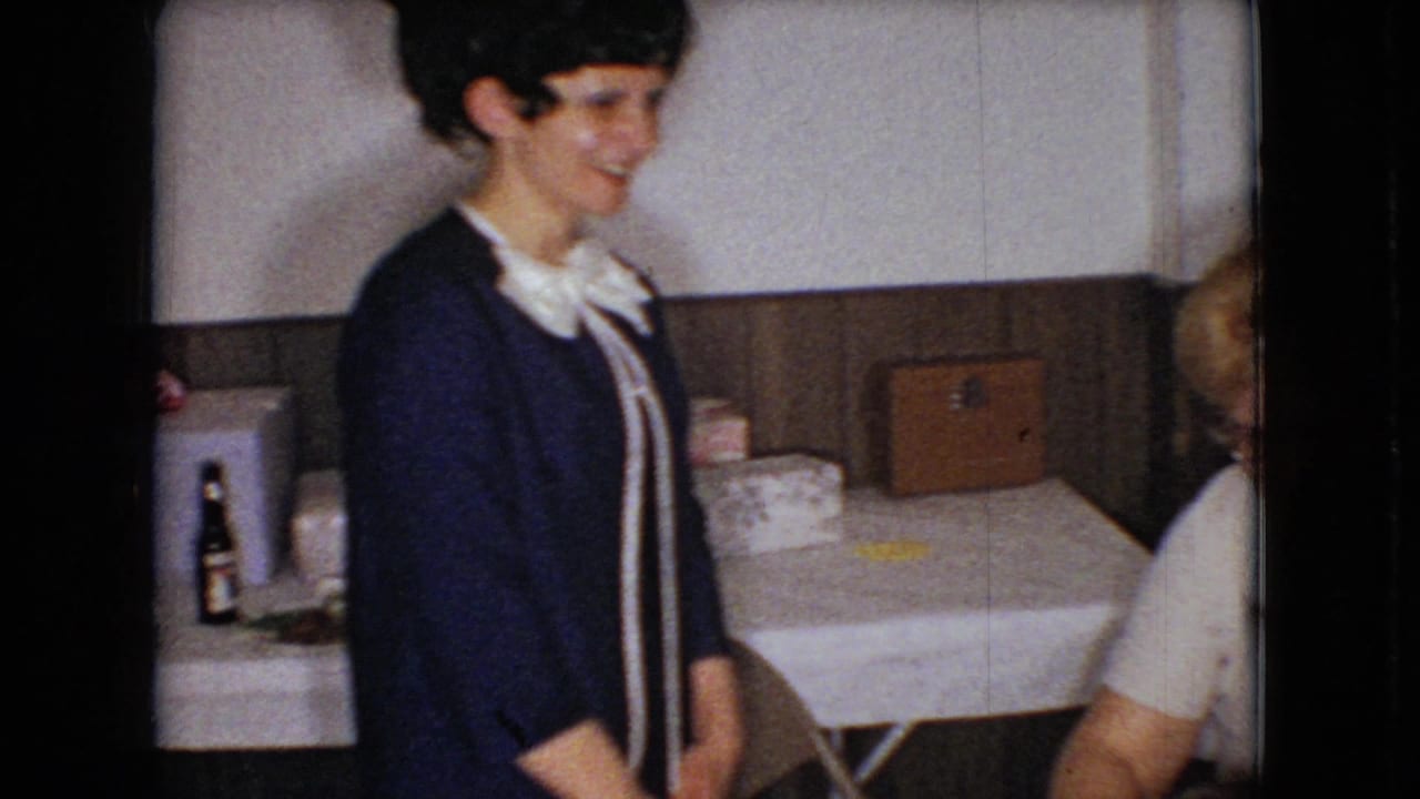 In supplied 1966 Ohio 8mm home-movie footage, a woman in a dark blue dress with a white bow collar smiles near a white tablecloth and wood-paneled wall.