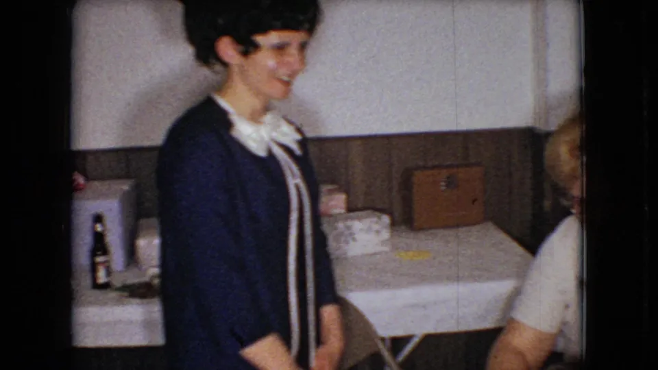 In supplied 1966 Ohio 8mm home-movie footage, a woman in a dark blue dress with a white bow collar smiles near a white tablecloth and wood-paneled wall.