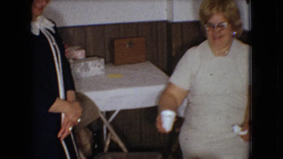 In supplied 1966 Ohio 8mm footage, a woman in glasses and a light dress is seen beside the table while the standing woman remains partly framed at left.