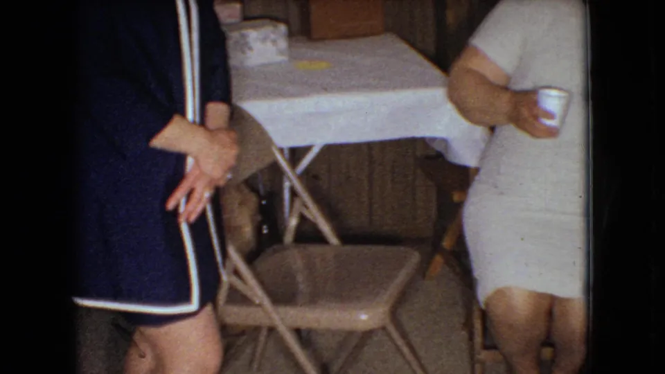 In supplied 1966 Ohio home-movie footage, a woman in a light dress holds a cup near folding chairs, a cloth-covered table, and wood-paneled walls.