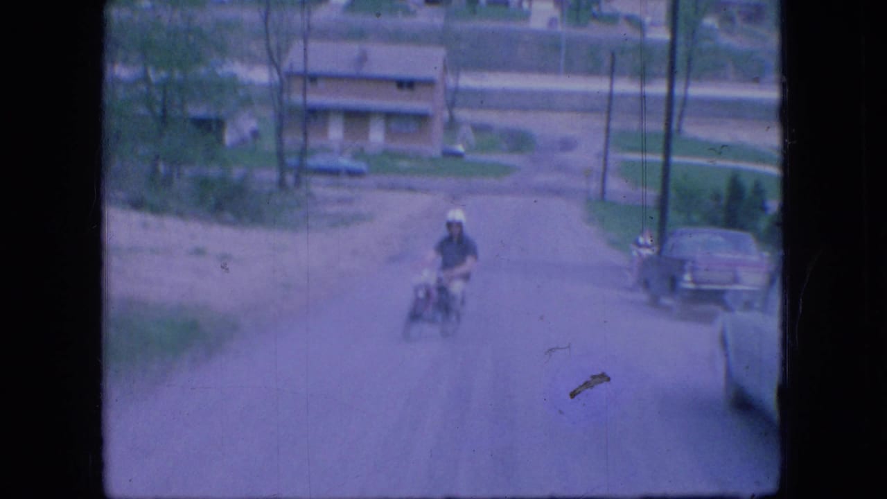 A helmeted adult man rides a small motorbike down a road in Ohio, with parked cars and low buildings visible in the background.