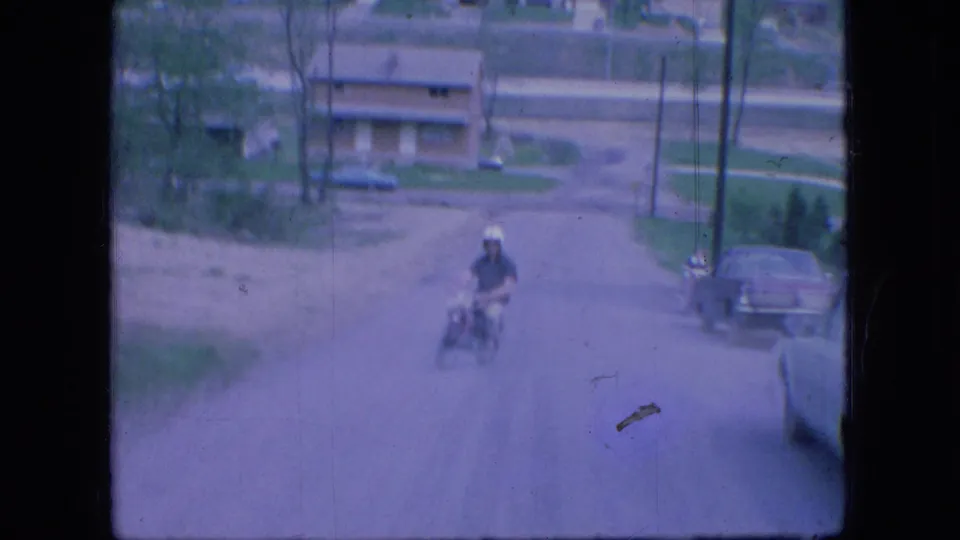 A helmeted adult man rides a small motorbike down a road in Ohio, with parked cars and low buildings visible in the background.