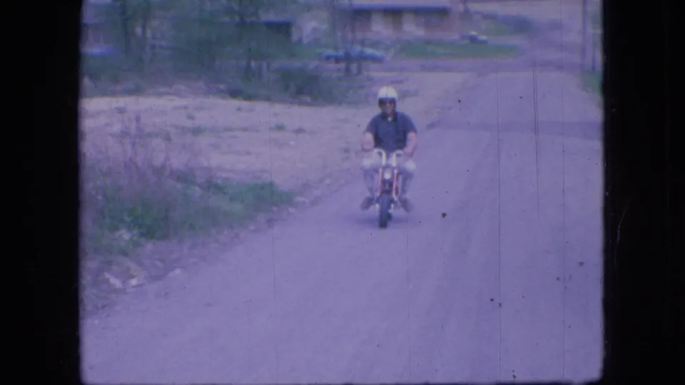 An adult rider in a white helmet and dark shirt approaches on a red mini bike along a rural or semi-rural Ohio road.