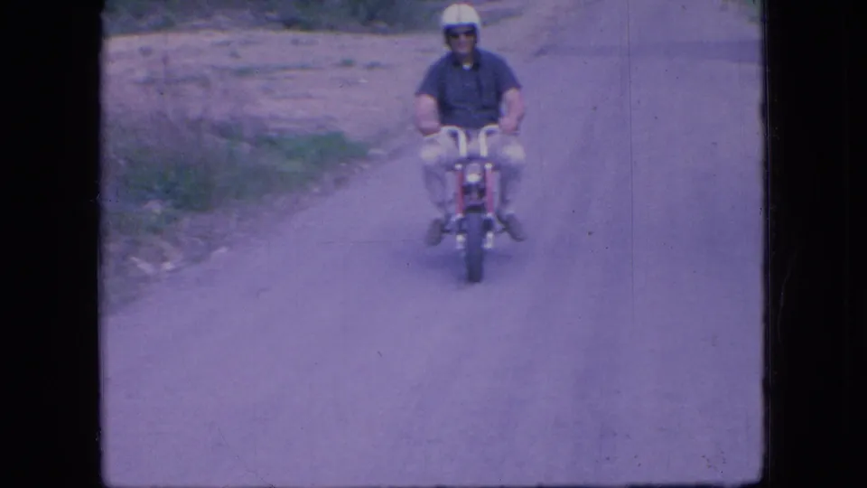 A helmeted adult man rides a red mini bike toward the camera on an Ohio road, framed with visible 8mm grain and soft color fading.