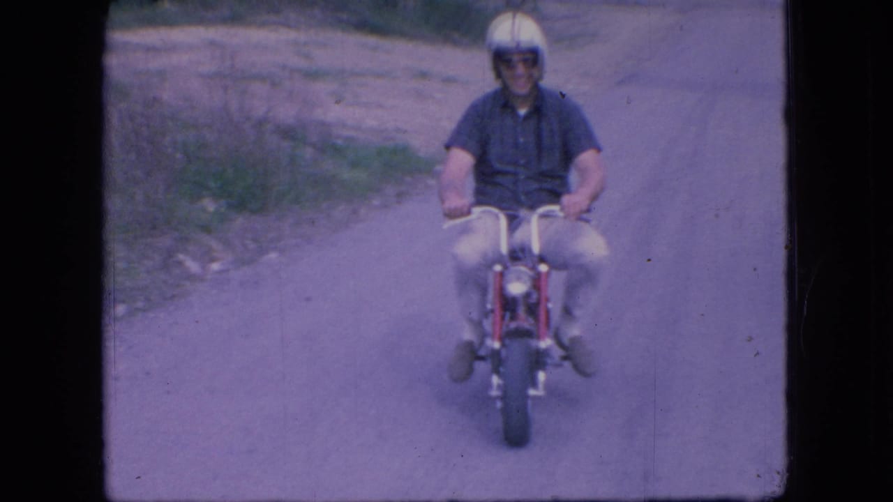 In 1966 Ohio home-movie footage, an adult man wearing a white helmet, dark sunglasses, blue shirt, and light pants rides a small red motorcycle toward the camera on a rural dirt road.