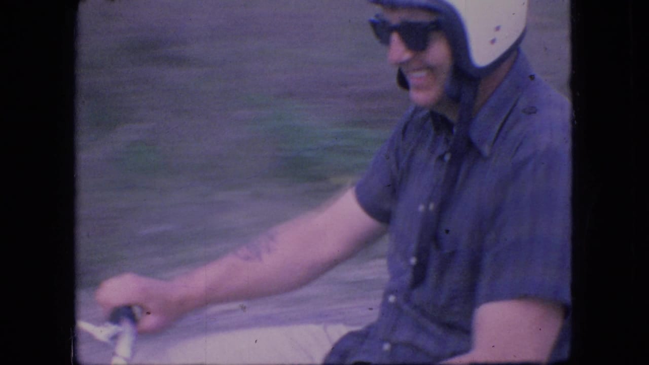 A close 1966 Ohio home-movie view shows the smiling rider in a white helmet and sunglasses passing the camera on a small motorcycle, with blurred roadside ground behind him.