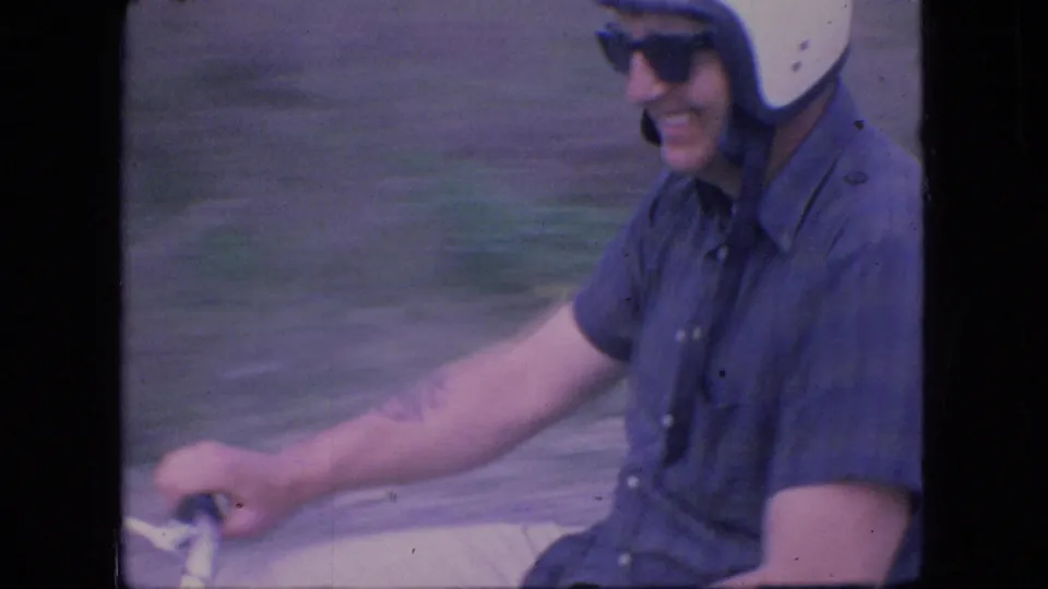 A close 1966 Ohio home-movie view shows the smiling rider in a white helmet and sunglasses passing the camera on a small motorcycle, with blurred roadside ground behind him.