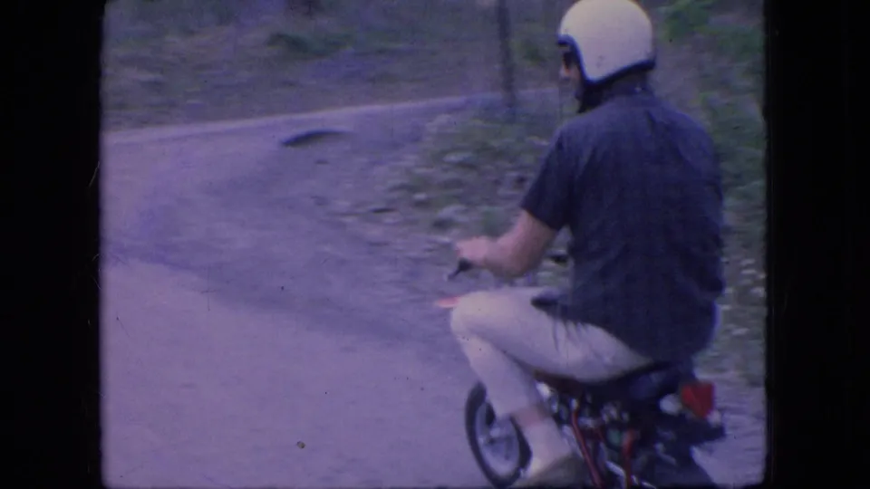 The helmeted man is seen from behind and slightly to the side as he rides the small red motorcycle along a curving dirt road in rural Ohio home-movie footage from 1966.
