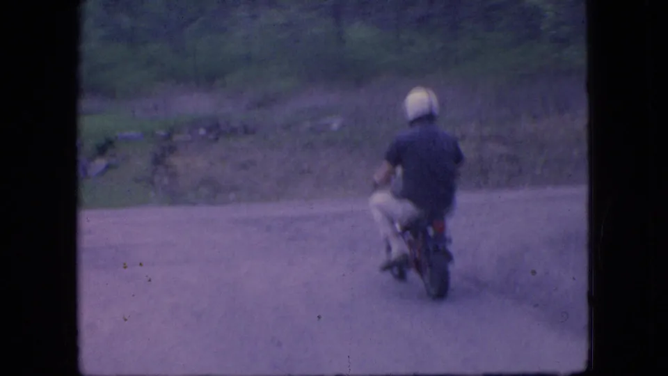 A distant rear view shows the man riding away on the small motorcycle down a rural dirt road, with brush and trees visible beyond the road in the 1966 Ohio footage.