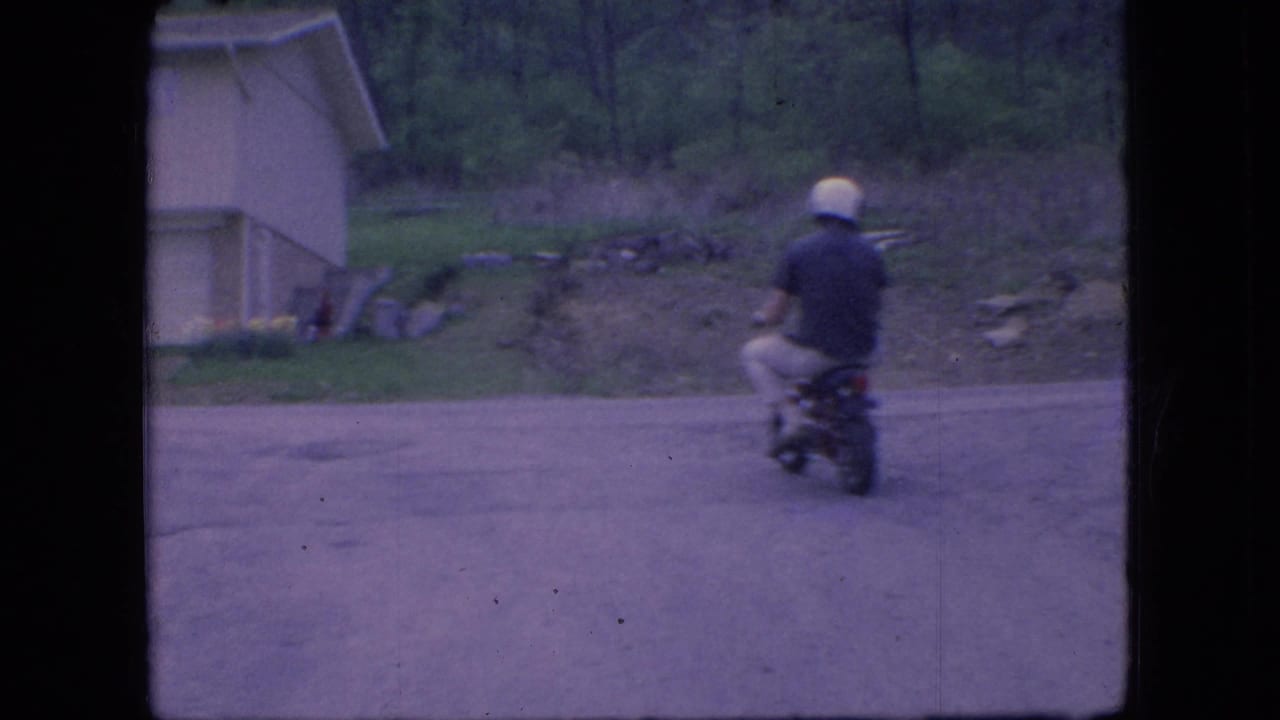 In 1966 Ohio home-movie footage, a helmeted rider on a small motorbike moves away from the camera on a quiet road beside a light-colored suburban house.