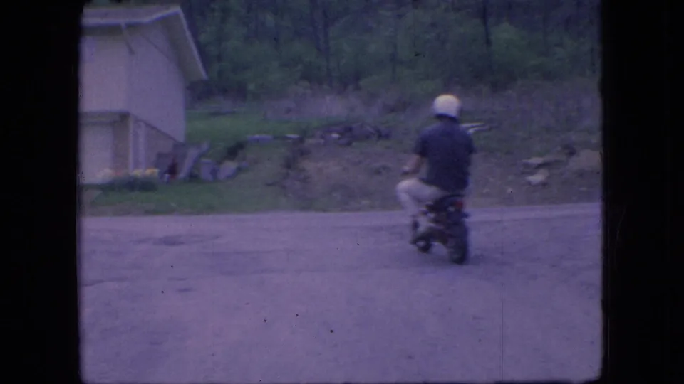 In 1966 Ohio home-movie footage, a helmeted rider on a small motorbike moves away from the camera on a quiet road beside a light-colored suburban house.