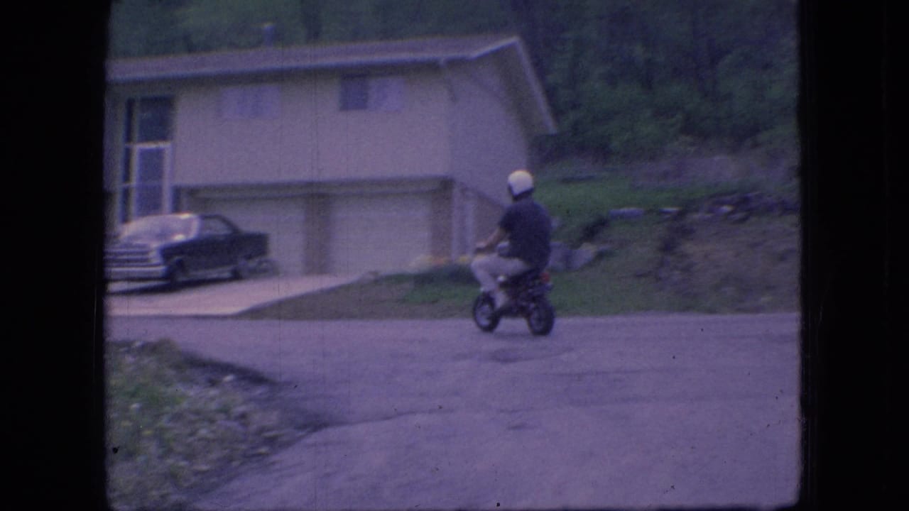 A helmeted rider on a mini bike passes a paved driveway and dark parked sedan in front of a 1960s suburban Ohio house.