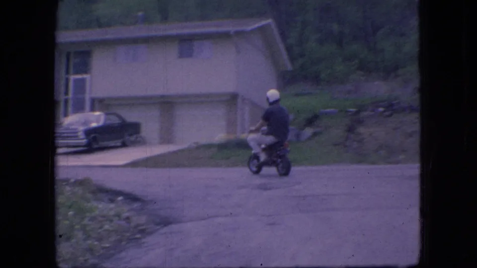 A helmeted rider on a mini bike passes a paved driveway and dark parked sedan in front of a 1960s suburban Ohio house.