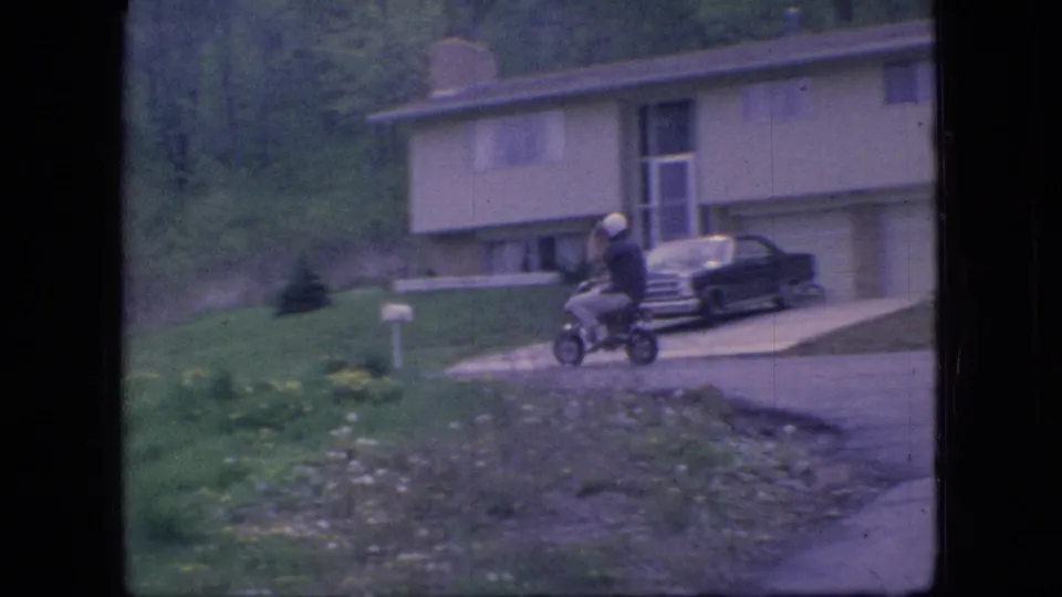The rider continues along the residential edge near a lawn and mailbox, with a split-level style house and parked sedan behind him in the Ohio home-movie frame.