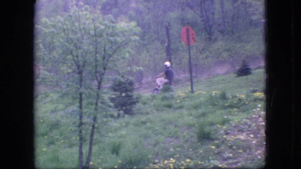 A helmeted mini bike rider appears farther from the camera in a grassy roadside area near a stop sign, with trees and spring or summer greenery around him.