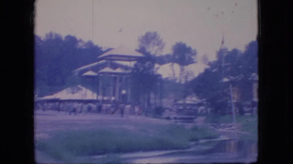 A soft, color-faded 1966 Ohio 8mm frame shows a covered pavilion beside grass and a creek or water edge, with trees, scattered people, tent-like structures, and parked automobiles in the background.