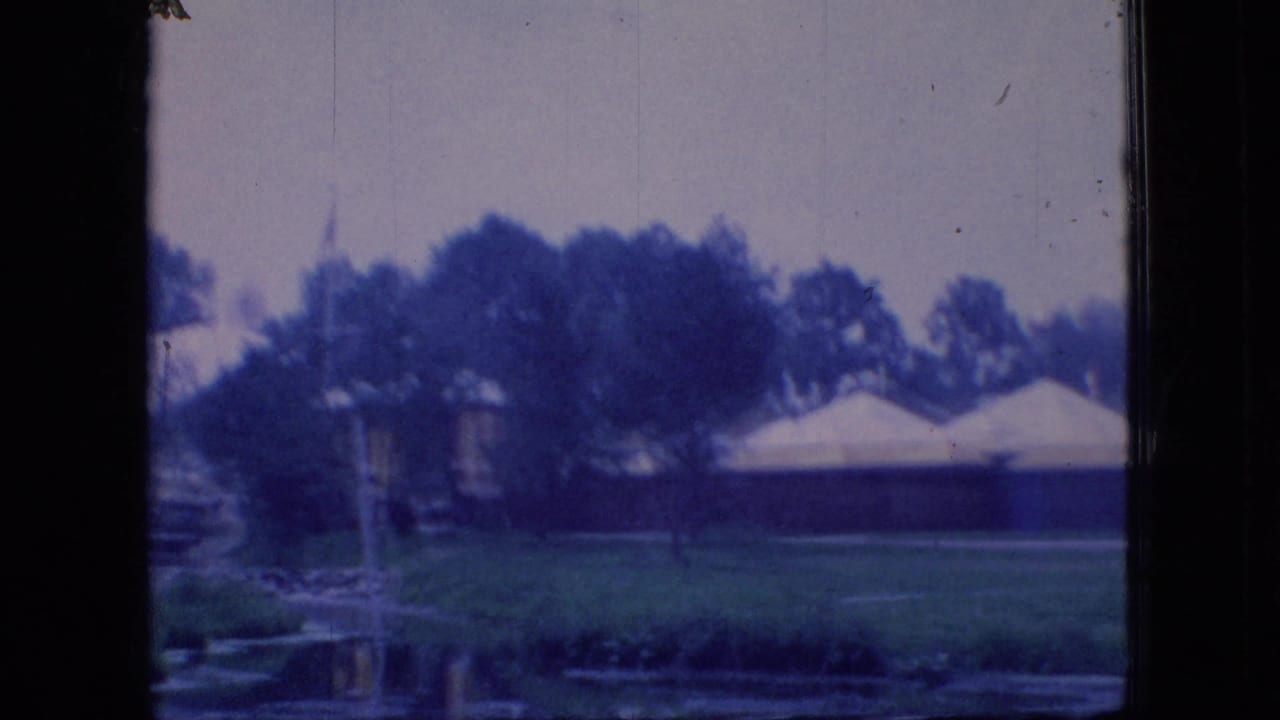 Blue-tinted 1966 Ohio home-movie frame of white tents, trees, lawn, and a pond edge seen from behind a dark vertical frame.