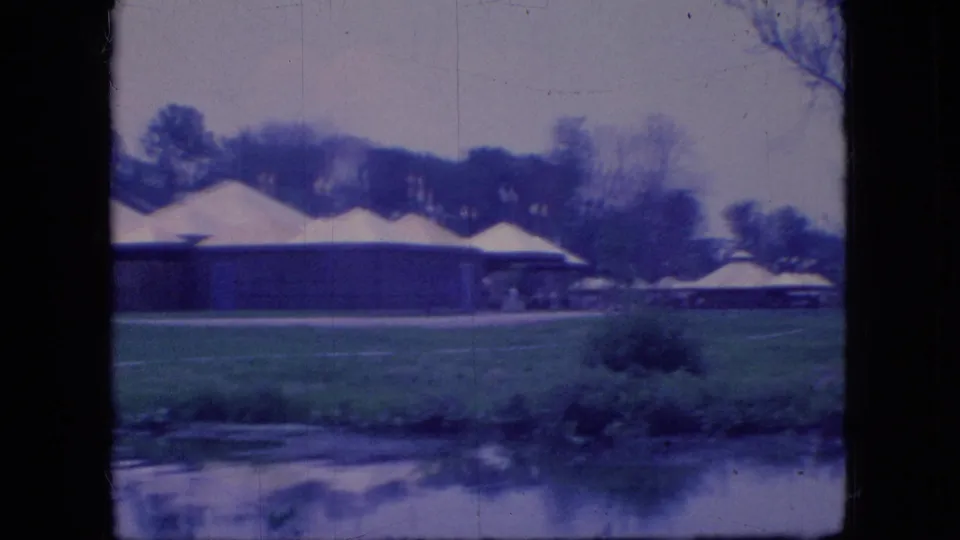 Blue-tinted 1966 Ohio 8mm frame with a low building, white tents, parked cars, lawn, shrubs, and pond water in the foreground.