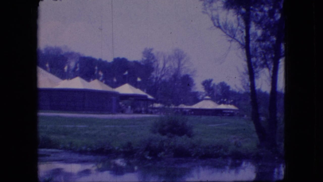1966 Ohio 8mm frame showing white-roofed pavilions or shelters across grass near calm water, with bare trees and shoreline reflections.