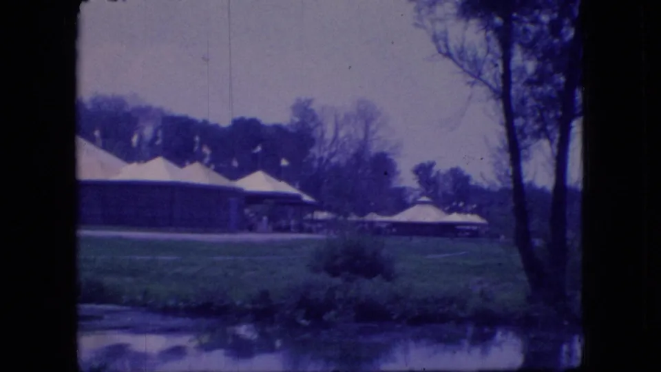 1966 Ohio 8mm frame showing white-roofed pavilions or shelters across grass near calm water, with bare trees and shoreline reflections.