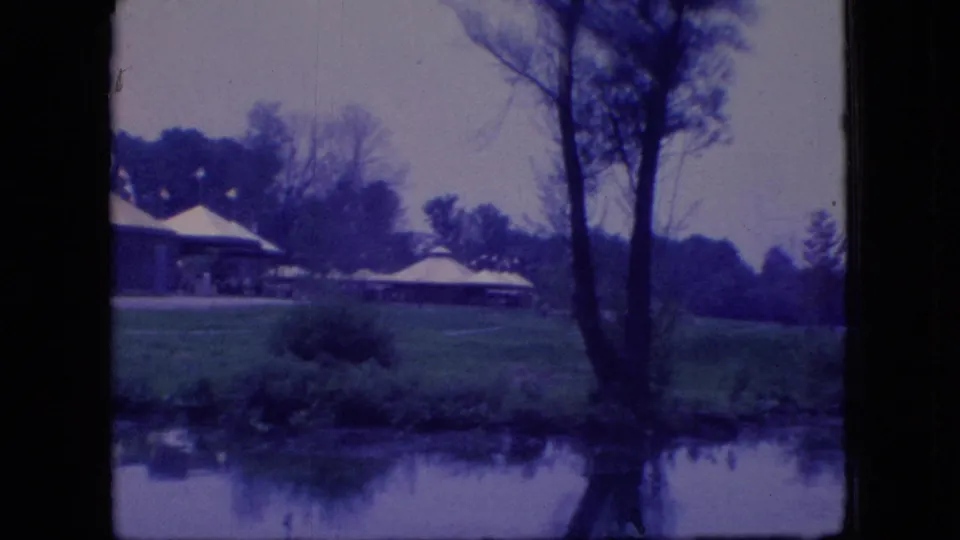 1966 Ohio home-movie frame with lakeside park shelters, open grass, trees, shrubs, and soft reflections in calm water.