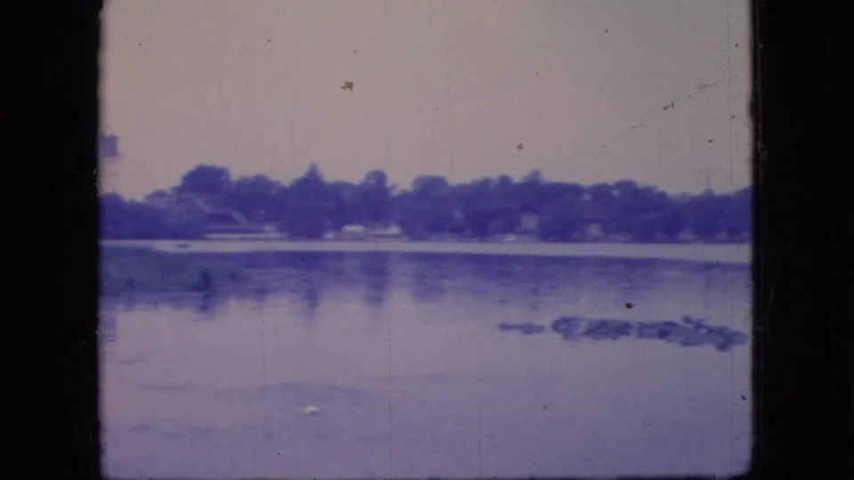 1966 Ohio lakeside frame showing still water, a far treeline, small distant structures or shelters, and patches of floating vegetation near the right foreground.