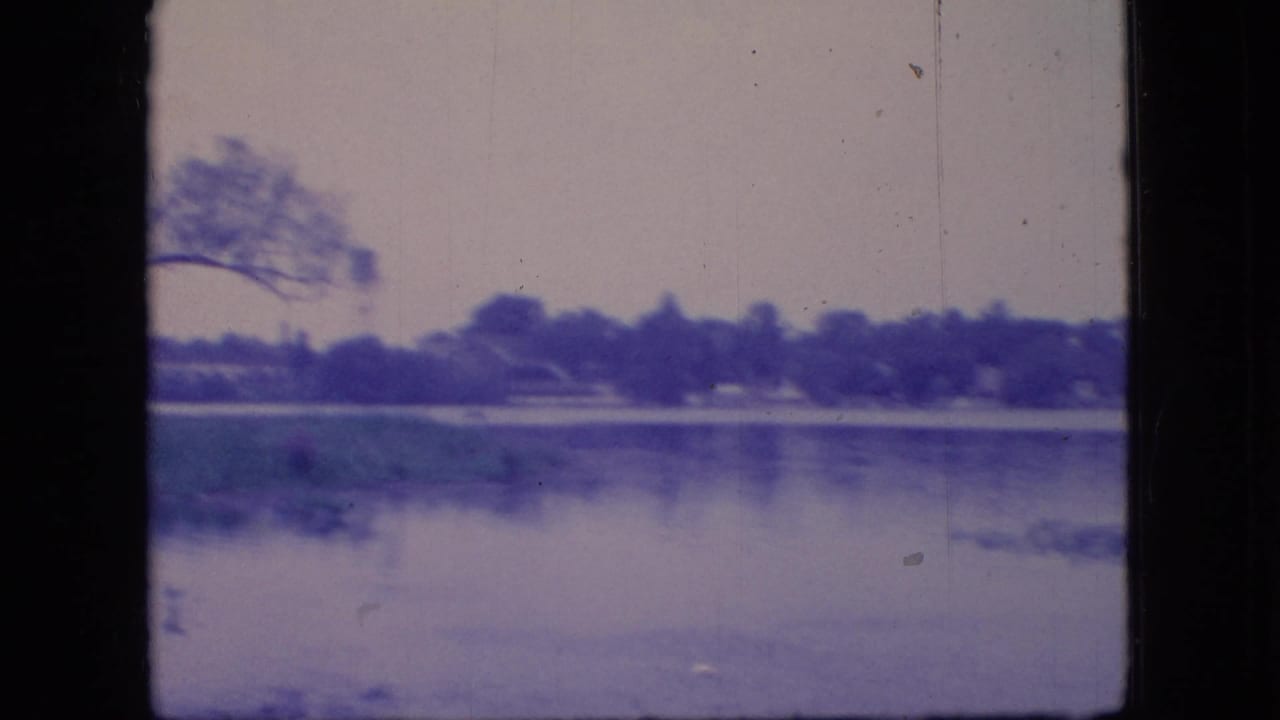 1966 Ohio 8mm frame showing a calm lake surface, distant treeline, and low shoreline vegetation with aged blue color cast.