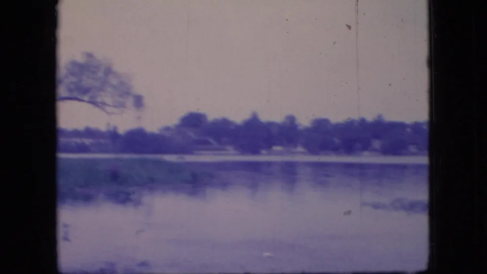 1966 Ohio 8mm frame showing a calm lake surface, distant treeline, and low shoreline vegetation with aged blue color cast.