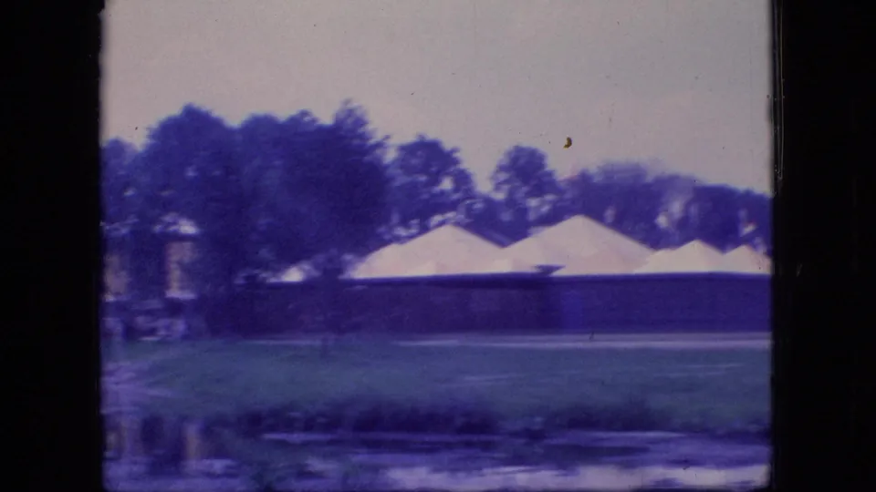 1966 Ohio park frame showing low pavilion structures with pale roof sections, trees, grass, and a narrow band of lake water in the foreground.